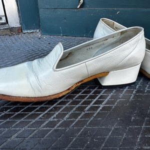Pu&ograve; includere: Un paio di scarpe bianche in pelle da infilare con un tacco basso a blocco. Le scarpe hanno una punta leggermente a punta e un design semplice.