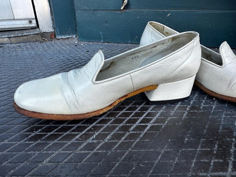 Pu&ograve; includere: Un paio di scarpe bianche in pelle da infilare con un tacco basso a blocco. Le scarpe hanno una punta leggermente a punta e un design semplice.