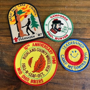 Puede incluir: Una colección de parches bordados vintage. Los parches presentan varios diseños, incluyendo un leñador, una carita sonriente y texto como "Great Salt Lake Council" y "Camp Metamora 1971". Los parches son de varios colores.