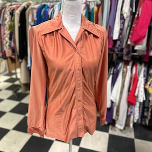 May include: A long-sleeved, button-down shirt in a muted peach color. The shirt has a collared neckline and is displayed on a mannequin. The fabric appears to have a slight sheen. The shirt is part of a vintage clothing collection.