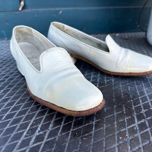 Pu&ograve; includere: Un paio di scarpe da barca in pelle bianca con punta arrotondata e tacco basso. Le scarpe hanno un aspetto leggermente usurato e sono realizzate in pelle morbida e flessibile.