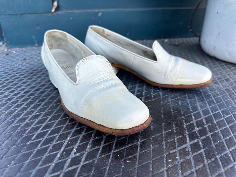 Pu&ograve; includere: Un paio di scarpe da barca in pelle bianca con punta arrotondata e tacco basso. Le scarpe hanno un aspetto leggermente usurato e sono realizzate in pelle morbida e flessibile.