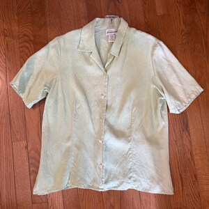 Puede incluir: Una camisa verde claro de manga corta con botones. La camisa tiene cuello y est&aacute; hecha de una tela ligera. Se muestra plana sobre una superficie de madera.