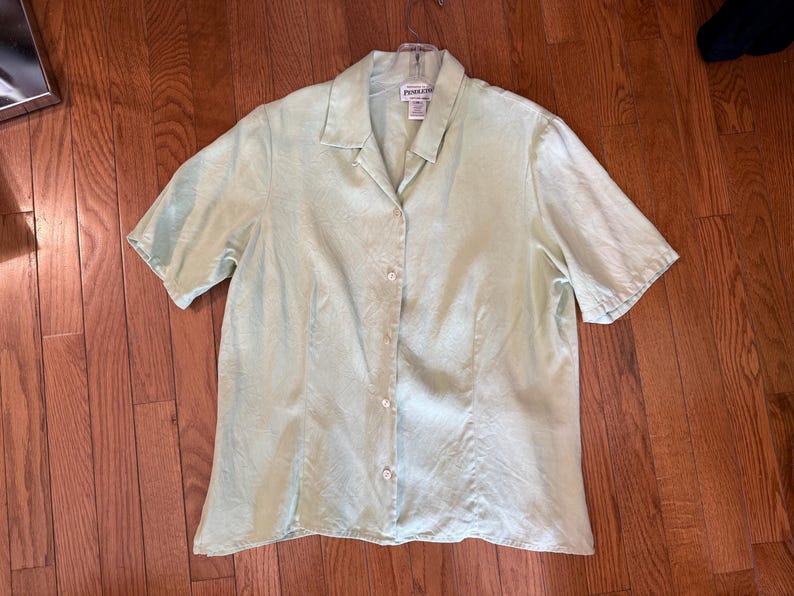 Puede incluir: Una camisa verde claro de manga corta con botones. La camisa tiene cuello y est&aacute; hecha de una tela ligera. Se muestra plana sobre una superficie de madera.