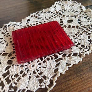 May include: A rectangular, translucent red object with vertical ridges, possibly a vintage accessory, rests on a cream-colored, openwork lace doily. The doily is placed on a dark wooden surface, creating a contrast of textures and colors.