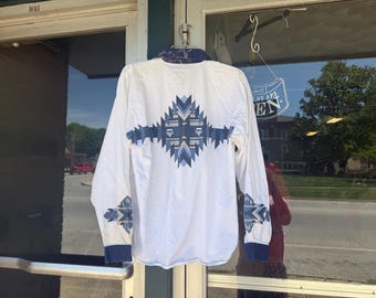 May include: White long-sleeve shirt with a geometric design in shades of blue on the back, cuffs, and collar. The shirt is hanging in front of a glass door, showcasing the back of the garment.