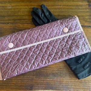 May include: A mauve quilted rectangular case with a diamond pattern, light pink trim, and two circular snap buttons, positioned over a pair of black gloves that feature small decorative bows on the wrist area of each glove.