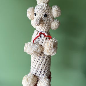 May include: A handmade, off-white crochet poodle toy with fluffy pom-pom accents. The poodle has black button eyes and a red ribbon collar. The toy is held against a green background.