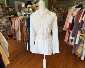 May include: A white long-sleeve blouse with embroidered detailing on the bodice and a woven belt. The blouse is button-down and has a collared neckline. The mannequin is displayed in a vintage clothing store, with racks of clothing in the background.