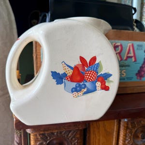 May include: White ceramic jug with a large handle and a fruit design. The jug features a blue bowl with red, blue, and white fruit illustrations. The jug is on a wooden surface.
