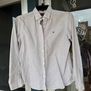 May include: A button-down, long-sleeve shirt with a striped pattern in shades of white and gray. The shirt has a button-down collar and a small logo on the chest. The shirt is hanging on a black hanger.