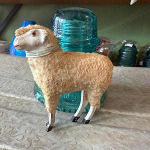 May include: A vintage sheep figurine with a tan body, white face, and black hooves. The sheep has a cream-colored collar and red accents on its face. It stands on a textured surface with a turquoise glass insulator in the background.