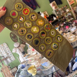 Op de afbeelding: Olijfgroene sjerp met geborduurde patches. Elke patch heeft een ander ontwerp, waaronder een hart, kruis en gereedschap. De patches zijn cirkelvormig met een gouden rand. De sjerp wordt door een persoon vastgehouden.