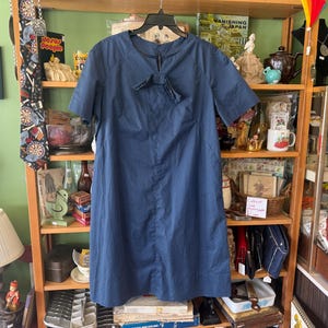 May include: A vintage navy blue dress with a white polka dot pattern. The dress features short sleeves and a bow detail at the neckline. It hangs on a black hanger against a wooden shelf filled with various items.