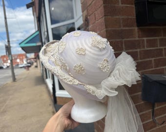 May include: A vintage white hat with delicate lace trim and floral embellishments. The hat features a rounded crown and a wide brim, with a sheer veil cascading down the back. A classic accessory for special occasions.