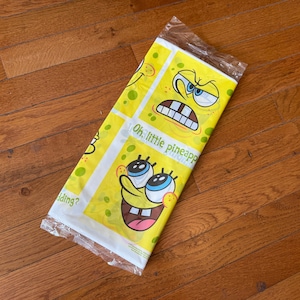 May include: A yellow plastic bag featuring SpongeBob SquarePants with various facial expressions. The bag has the text "Oh, little pineapple" and is sealed at the top. The bag is lying on a wooden floor.