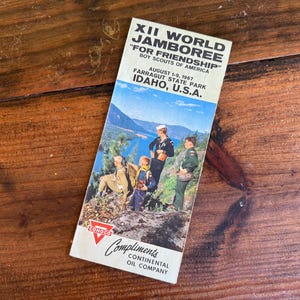 May include: A vintage brochure for the XII World Jamboree, a Boy Scouts of America event held in Idaho, USA, in 1967. The brochure features a scenic image of scouts and a sailor overlooking a lake and mountains, with the Conoco logo.