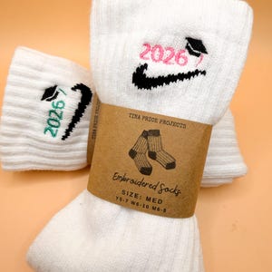 May include: White embroidered socks with the year "2026" in pink and green, along with a graduation cap design. The socks are packaged with a brown paper label that reads "Embroidered Socks" and "TINA PRICE PROJECTS".