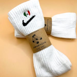 Puede incluir: Calcetines blancos bordados con un swoosh Nike negro y un corazón verde, blanco y rojo con una pequeña bandera mexicana en su interior. Los calcetines están etiquetados como "Embroidered Socks" con la talla "MED" y "7-7 W 8-10 MS 8-8".