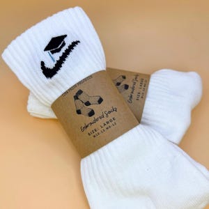 以下が含まれることがあります： 黒い刺繍の卒業キャップとスウッシュロゴが付いた白いスポーツソックス。ソックスは「Embroidered Socks」と「SIZE: LARGE W10-13 M8-12」と書かれた茶色の紙バンドで包まれています。「TINA PRICE PROJECTS」の文字も表示されています。