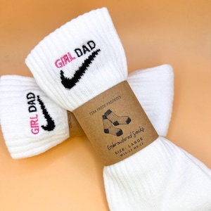 May include: White athletic socks with the text "GIRL DAD" embroidered in pink and black. The socks feature a black swoosh design. A brown paper band around the socks reads "Embroidered Socks" and "SIZE: LARGE".