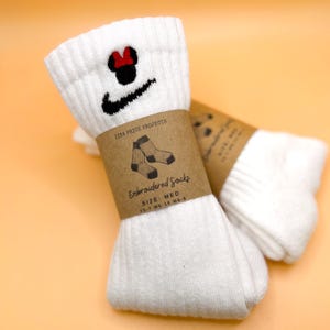 以下が含まれることがあります： 黒いナイキのロゴとミニーマウスのデザインが刺繍された白い靴下。靴下は「Embroidered Socks」と「Size: Med」と書かれた茶色の紙バンドで包まれています。