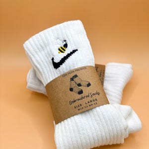 May include: White ribbed socks with a black and yellow embroidered bee design and a black checkmark. The socks are wrapped with a brown paper band that reads "Embroidered Socks" and "Size: Large W10-13 M8-12".