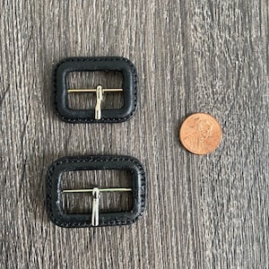 Puede incluir: Dos hebillas de cinturón de cuero negro con cierres de metal plateado. Las hebillas son de diferentes tamaños.