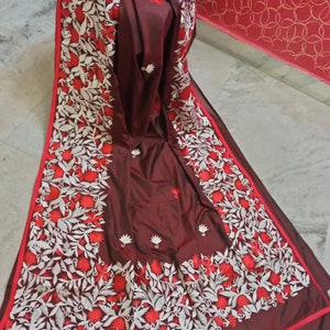 Pu&ograve; includere: Un sari di seta marrone con ricami bianchi e rossi. Il ricamo presenta disegni floreali e motivi intricati.