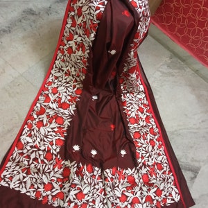 Pu&ograve; includere: Un sari di seta marrone con ricami bianchi e rossi. Il ricamo presenta un motivo floreale con foglie e fiori.