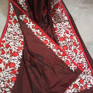 Pu&ograve; includere: Un sari di seta marrone con ricami bianchi e rossi. Il ricamo presenta un motivo floreale con foglie e fiori.