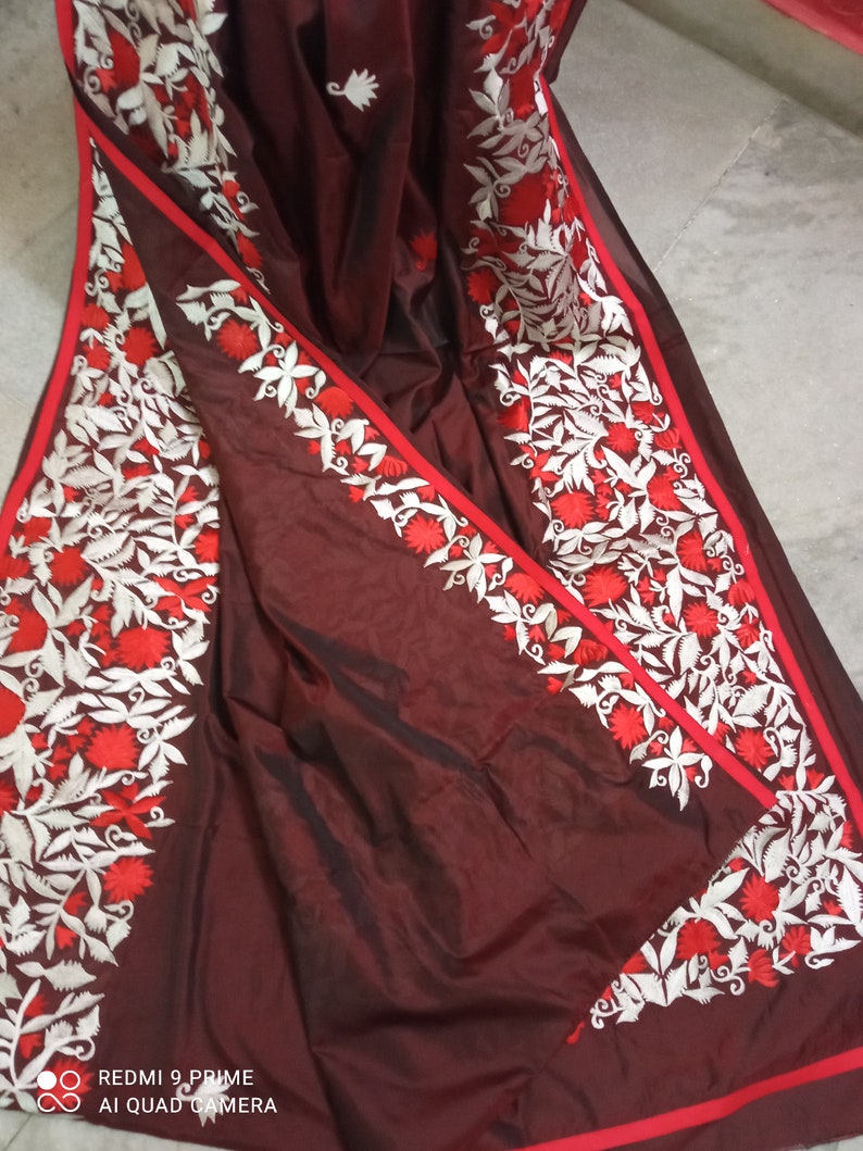 Pu&ograve; includere: Un sari di seta marrone con ricami bianchi e rossi. Il ricamo presenta un motivo floreale con foglie e fiori.