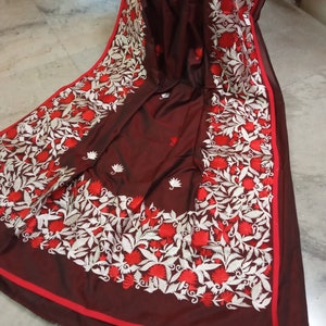 Pu&ograve; includere: Un sari di seta marrone scuro con ricami bianchi e rossi. Il ricamo presenta un motivo floreale con foglie e fiori.