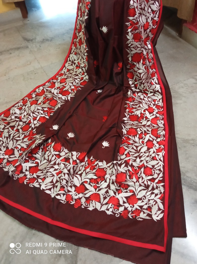 Pu&ograve; includere: Un sari di seta marrone scuro con ricami bianchi e rossi. Il ricamo presenta un motivo floreale con foglie e fiori.