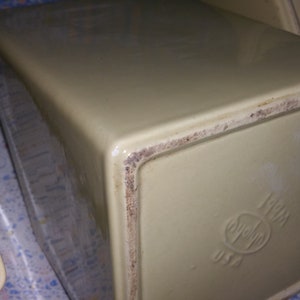 Peut inclure: Gros plan du fond d'un pot en c&eacute;ramique de couleur cr&egrave;me avec un bord brun. Le pot a un logo avec un c&oelig;ur et le texte "USA" imprim&eacute; au fond.