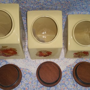 Peut inclure: Ensemble de trois pots en c&eacute;ramique avec couvercles en bois. Les pots sont de couleur cr&egrave;me avec un motif floral et pr&eacute;sentent une ouverture ronde en haut. Les couvercles sont en bois brun.
