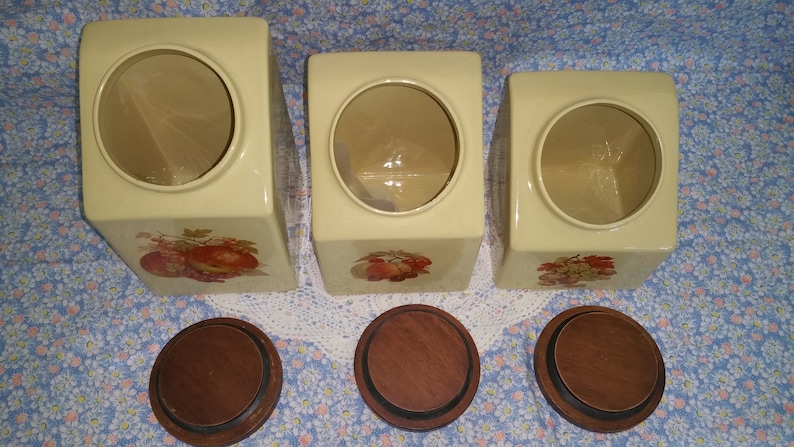 Peut inclure: Ensemble de trois pots en c&eacute;ramique avec couvercles en bois. Les pots sont de couleur cr&egrave;me avec un motif floral et pr&eacute;sentent une ouverture ronde en haut. Les couvercles sont en bois brun.