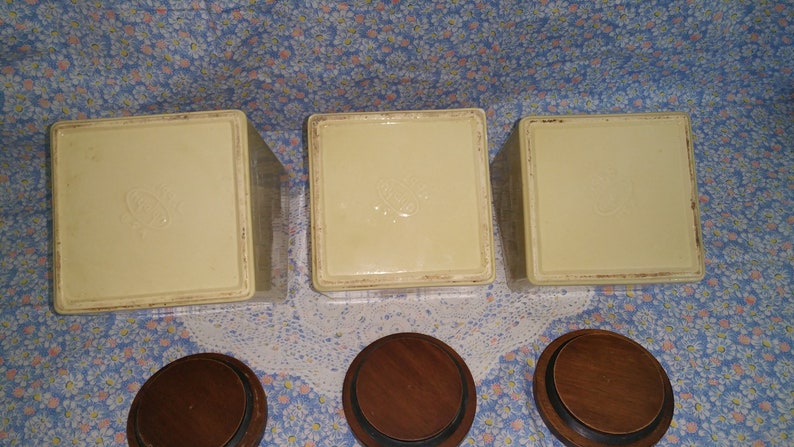 Peut inclure: Trois pots en c&eacute;ramique blanche avec couvercles. Les pots ont un motif en relief sur le dessus et sont pos&eacute;s sur une surface &agrave; motifs floraux. Les couvercles sont en bois et ont une finition brun fonc&eacute;.