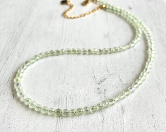 May include: A delicate necklace featuring small, faceted, pale green beads. The beads are strung together to form a necklace with a gold-colored chain and clasp. The necklace is displayed on a white wooden surface.