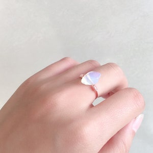 May include: A delicate ring featuring a white iridescent gemstone wrapped in rose gold wire.