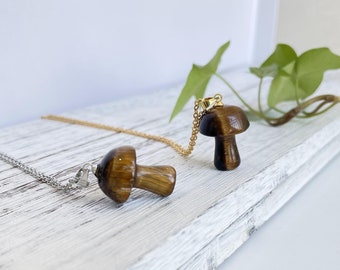 Puede incluir: Dos collares de oro con colgantes de hongo de ojo de tigre. Los collares están sobre una superficie de madera blanca.