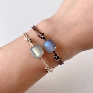 May include: Two bracelets with brown and beige cords, each featuring a square-shaped labradorite gemstone bead. The beads have a light blue and gray iridescent sheen.