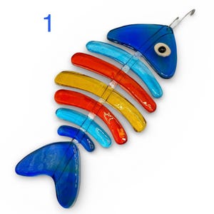 May include: A colorful glass fish skeleton ornament. The ornament features a blue head and tail, with a spine made of blue, red, and yellow curved glass pieces. A small eye is visible on the head. A metal wire is attached for hanging.