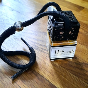 May include: A black car air freshener with a black and gold decorative cage. The air freshener is attached to a black string with a silver bead. The label on the air freshener reads "JT-Scents handmade in Scotland."