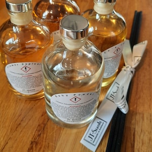 May include: Four glass bottles with silver lids and labels containing scented oil. The labels have warning signs and text that reads "Flower Explosion", "Baby Powder", and "Jo's Scent". The bottles are sitting on a wooden surface with black diffuser sticks.