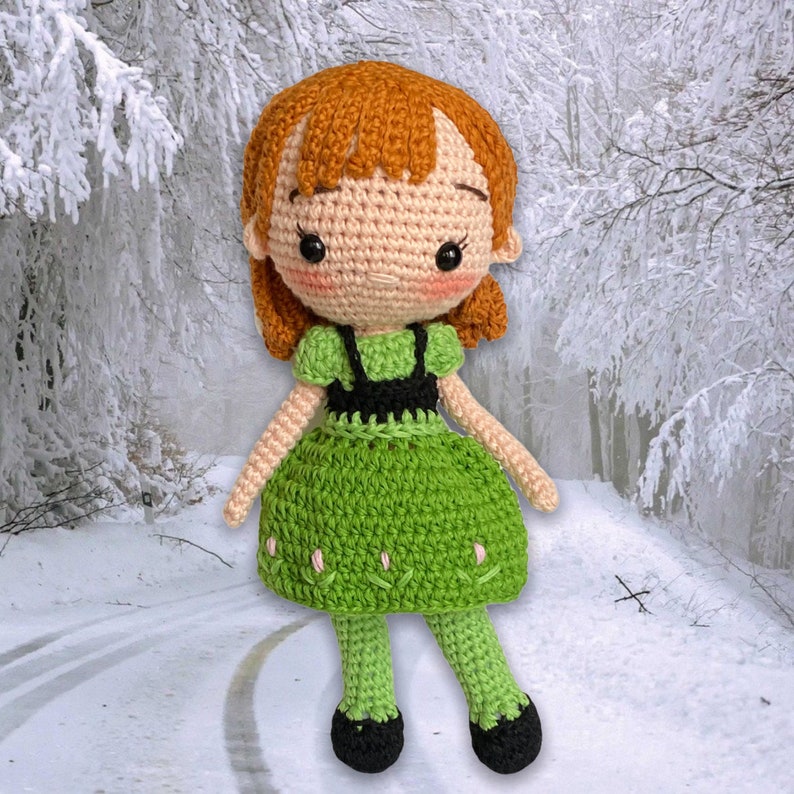 May include: A crocheted doll with ginger hair, rosy cheeks, and black eyes. The doll is dressed in a green dress with a black bodice and green legs with black shoes. The background shows a snowy winter scene.