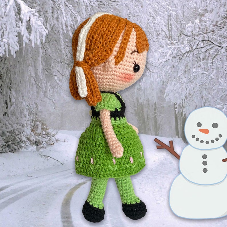 May include: A handmade crochet doll with ginger hair, wearing a green dress with black detailing. The doll has rosy cheeks and is set in a snowy scene next to a snowman.