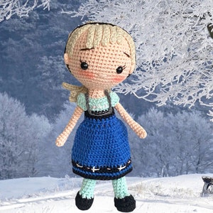 May include: A crocheted doll with a fair complexion, blonde hair, and rosy cheeks. The doll is dressed in a blue dress with black and white details, a mint green top, and black shoes. The doll's arms and legs are poseable, and it has a gentle expression.