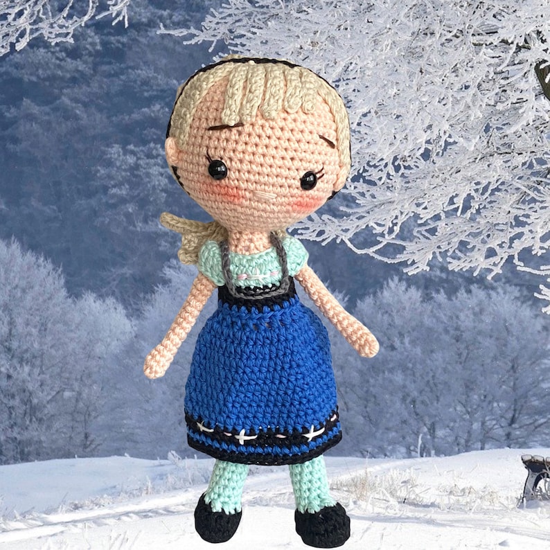 May include: A crocheted doll with a fair complexion, blonde hair, and rosy cheeks. The doll is dressed in a blue dress with black and white details, a mint green top, and black shoes. The doll's arms and legs are poseable, and it has a gentle expression.