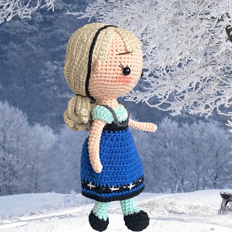May include: A crocheted doll with blonde hair, wearing a blue dress with black detailing, and black shoes. The doll has a black headband and rosy cheeks. The background shows a snowy winter scene.